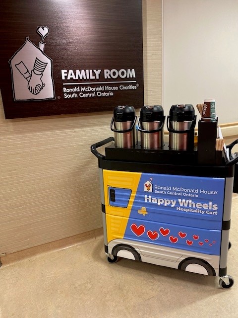 cart with fourwheels in front of signage that reads Ronald McDonald House Charities Family Room. CArt is wrapped in a cling that looks like a yellow school bus.